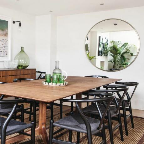 Трпезарија уређена у белој боји са дрвеним подом, дрвеним трпезаријским столом са старинским столицама и округлим огледалом. Зелени прибор и биљке. Западни Лондон дом Габи Палумбо и њеног мужа Метјуа