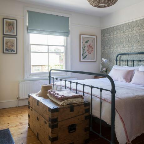 Bela spalnica s kovinsko bolniško posteljo, rožnata posteljnina, zavesa iz račjega jajca, tapeta William Morris, lesena skrinja