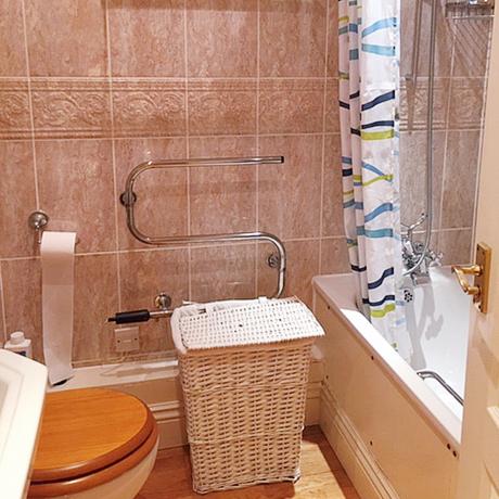 Zastaraná kúpeľňa s neutrálnymi dlaždicami a toaletnou doskou s dreveným efektom, sprchovací kút v bielej vani s farebným vzorovaným dekorom sprchového závesu