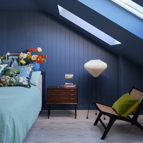 ไอเดียสีห้องนอนเล็ก ห้องนอนลอฟท์สีกรมกับผนังและเพดานสีฟ้า โต๊ะข้างวินเทจขนาดเล็ก โคมไฟตั้งพื้น เก้าอี้ ผ้าปูที่นอนสีฟ้าซีด หมอนอิงลาย แจกันใส่ดอกป๊อปปี้