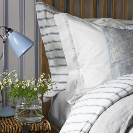 Lova su pagalvėmis, mėlynai baltai dryžuota patalynė ir gėlėmis ant naktinio staliuko