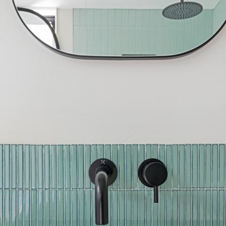 Un prim-plan cu accesorii pentru robinete de baie de culoare neagră mat, montate pe plăci de mozaic galben verde, cu oglindă dreptunghiulară moale în fundal, care afișează dispozitivul de duș în reflex