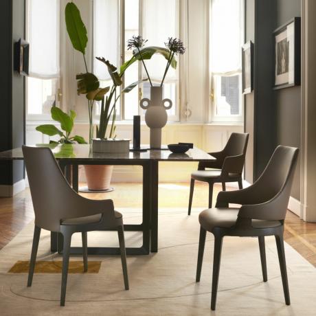 мала елегантна трпезарија са модерним смеђим столицама, квадратним столом, тепихом, биљкама, вазама, уметничким делима, смеђим зидовима, великим прозором у позадини