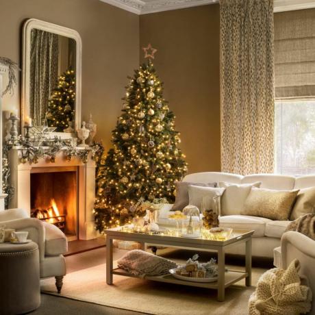 Rotā Ziemassvētku eglīte lauku stilā brūnā viesistabā, balts dīvāns, spilveni, kamīns, iekurts ugunskurs, dekorēts kamīns, liels spogulis, kafijas galdiņš.