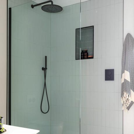 Un prim plan al unei cabine de duș cu cap de duș negru mat și accesorii cu colț care conține produse de baie în recipiente din sticlă maro