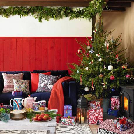 Kalėdų dekoravimo idėjos, žalumynų girliandos ant lubų, mėlyna sofa priešais raudonai nudažytas dailylentes, pagalvėlės su gėlių raštais, krūva dovanų po papuošta eglute