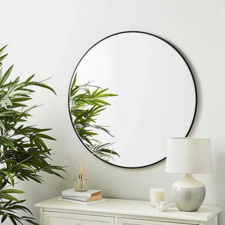 مرآة دنلم مستديرة بإطار أسود مثبتة على الحائط فوق خزانة جانبية بيضاء بجوار نبات كبير مع كتب ومصباح على الجانب