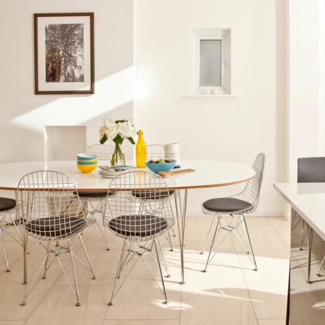 біла їдальня з овальним столом, сучасні дротяні стільці, жовто-бірюзовий посуд, витвори мистецтва, ламінат із світлого дерева