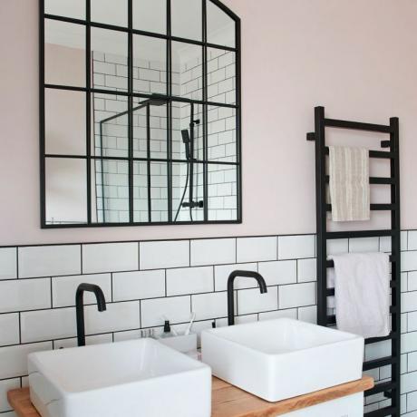 Розовая и монохромная ванная комната с двойной раковиной на деревянном умывальнике, черная вешалка для полотенец и зеркало на оконном стекле