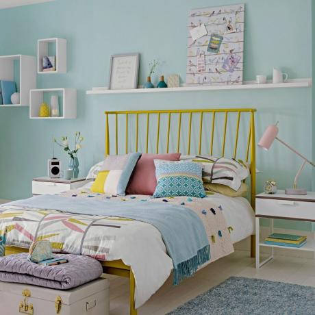 vaaleansininen makuuhuone, jossa valkoiset hyllyt ja keltainen sängynrunko vaaleanpunaisilla ja sinisillä vuodevaatteilla