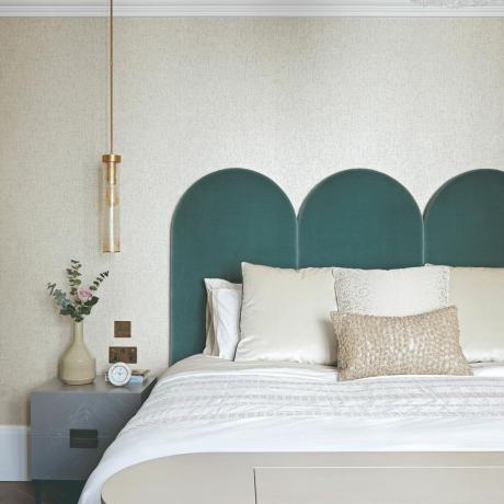 Sänky, jossa lämpimänharmaat tyynyt ja harmaa pääty, laivastonsininen sängynpääty, lämpimänharmaa seinä ja yöpöytä kasveilla