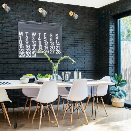 Jedilni prostor z belo mizo in stoli ter črno pobarvano izpostavljeno opečno steno