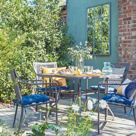 Záhrada so súpravou stolov a stoličiek a záhradným zrkadlom na tehlovej stene.
