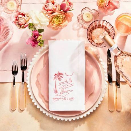 Pink bordlandskab opsat med tallerkener, bestik og en broderet serviet.
