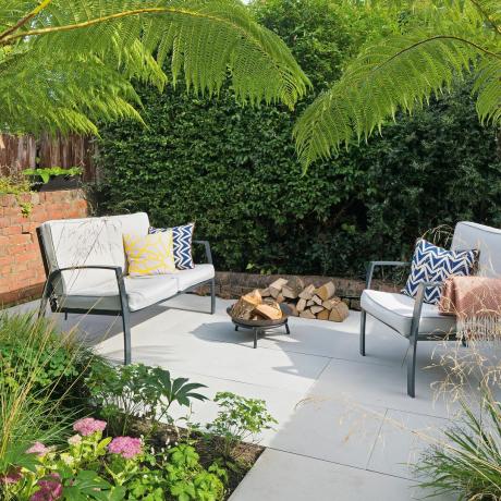 Brukowane patio ze współczesną sofą ogrodową i fotelem obok małego paleniska z wiszącymi palmami