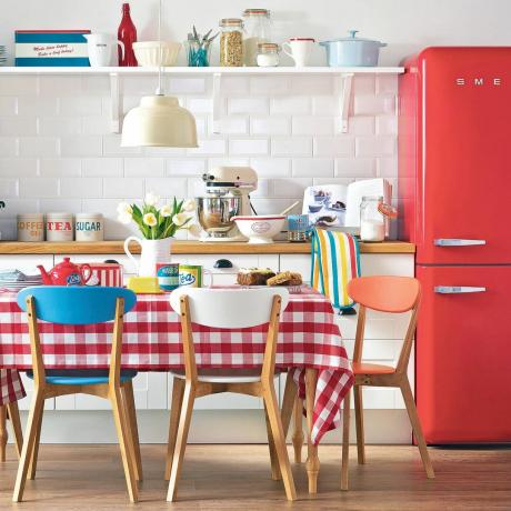 virtuvė su mediniu valgomuoju stalu su raudona gingham staltiese, baltomis plytelėmis ir raudonu Smeg šaldytuvu