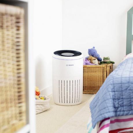 Un purificador de aire Bosch blanco en una habitación infantil al lado de una cama con ropa de cama a rayas