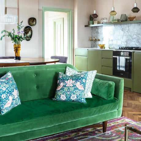 Žalia sofa su pagalvėlėmis tamsaus medžio svetainėje ir virtuvėje