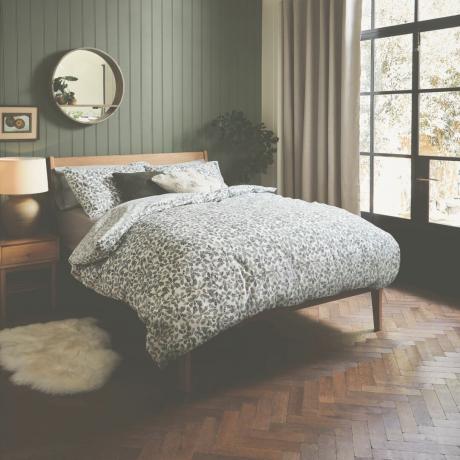 rustikálna spálňa so zelenou stenou na pero a drážku, parketová podlaha, krištáľové dvere smerom von, vzorovaná posteľná bielizeň, drevené zrkadlo, drevená posteľ a zodpovedajúci príručný stolík, koberec z ovčej kože