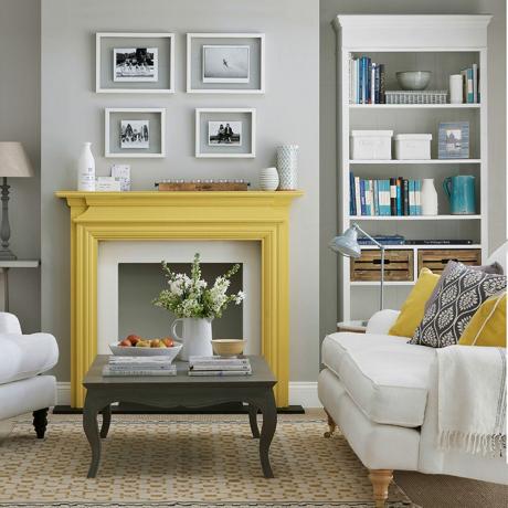 dzīvojamā istaba ar dzeltenu kamīna apkārtni, pelēkām sienām un baltu grāmatu skapi un krēmkrāsas krēslu