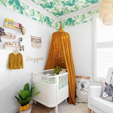 Weißes Kinderzimmer mit tapezierter Decke im Kaktusmuster und einem ockerfarbenen Baldachin über dem weißen Kinderbett