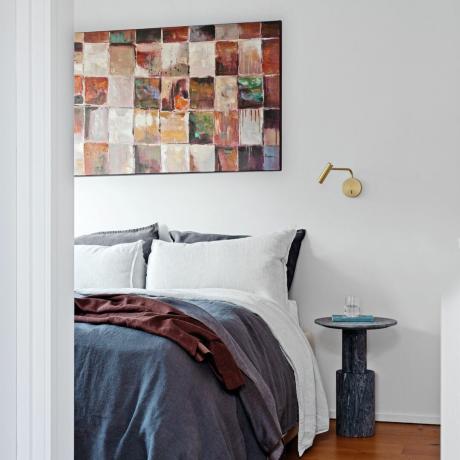 חדר שינה צבוע לבן עם אמנות קיר מעל המיטה ומצעים אפורים