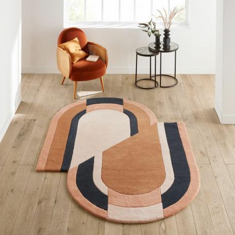 Oválný geo designový koberec v hnědé, krémové a zelené barvě na dřevěné podlaze