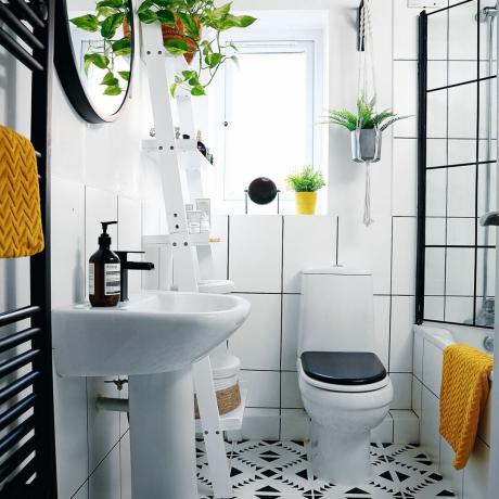 Cuarto de baño con azulejos blancos, estante de escalera blanco con plantas y espejo colgante circular
