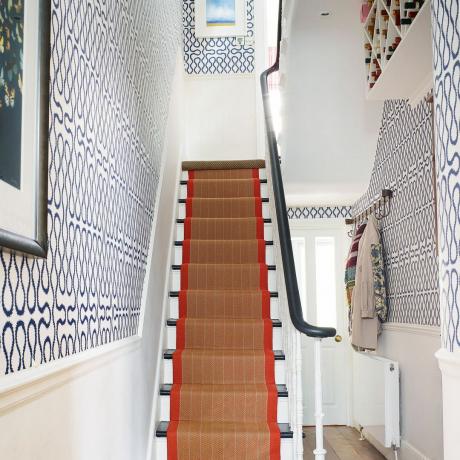 壁にジオ スタイルの大胆な壁紙が貼られた白い廊下と、赤い縁取りの天然床の階段ランナー