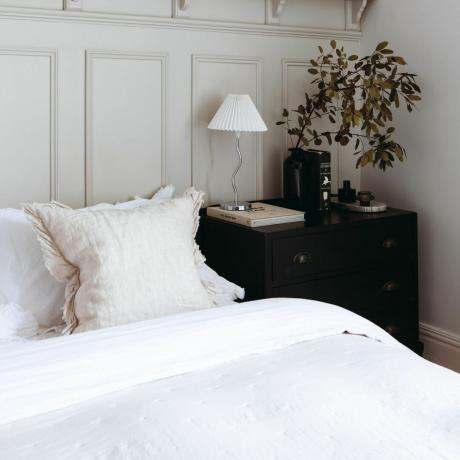 Obložená stěna ložnice, postel s bílým povlečením, černá noční komoda