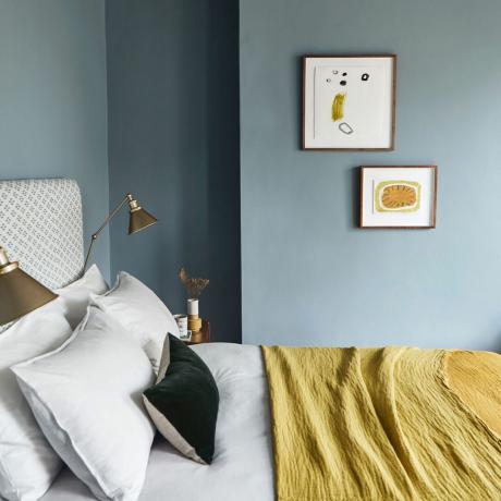 Quarto azul com manta de mostarda, roupa de cama branca, cabeceira estofada e arte na parede